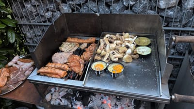 This image captures a close-up, eye-level view of a barbecue grill in Dissen am Teutoburger Wald, Germany. The grill is actively cooking a variety of foods. On the left side of the grill, several pieces of meat are being cooked over hot coals. These include what appear to be seasoned chicken pieces, pork chops, and possibly bacon-wrapped sausages arranged on a metal platter. On the right side of the grill, a stainless steel tray holds sautéed mushrooms and sliced eggplant. Two small metal rings contain fried eggs, with the yolks still runny. The background reveals a gabion wall filled with grey stones, characteristic of landscaping in the Teutoburg Forest region. Lush green foliage is visible to the left of the grill. The overall scene depicts an outdoor cooking event in a semi-urban or suburban setting.