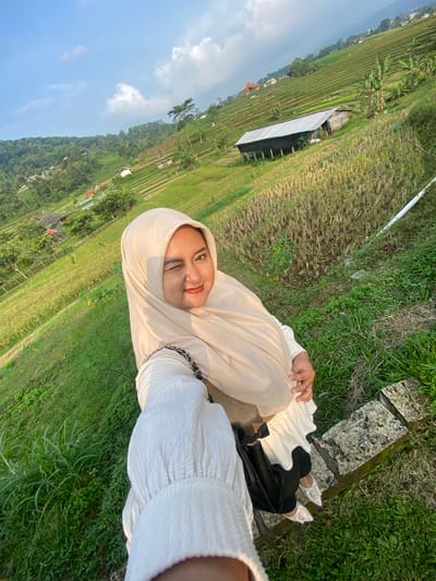 The image shows a person wearing a beige hijab and a white outfit, posing in front of a rural landscape. The person is smiling and winking at the camera. Behind the person is a lush, green terraced field. To the left of the terraced fields, various buildings and structures are partially visible. The image appears to be taken in a natural setting, likely in the city of Tamiajeng, Indonesia given the environment. The image has been rotated; the subject is lying or leaning.