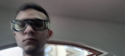 The image shows the head and shoulders of a young man wearing dark framed, mirrored sunglasses, in the front passenger seat of a car. He has short dark hair. The image appears to be taken by the driver from the front passenger seat. The only other discernible detail is a partially visible rear window, which indicates the car is moving.  The location is inside a car at the coordinates provided, which places the vehicle in Encarnación, Paraguay.