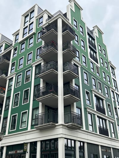This is a low-angle exterior shot of a modern, multi-story building with a distinctive stepped facade. The building features an emerald green exterior with white trim outlining the windows and structural elements. Numerous windows, many with black frames, are visible across the facade. Several balconies with black metal railings jut out from the building. The sky is overcast, suggesting a cloudy day.
On the right side of the building, a storefront is visible. A green and white sign with the text "BAGELS BEATIS" is prominently displayed, indicating a food establishment. The overall scene is urban, and the architectural style of the building suggests a contemporary design. There are no people clearly visible in the image. The composition and angle create a sense of grandeur and scale for the building.