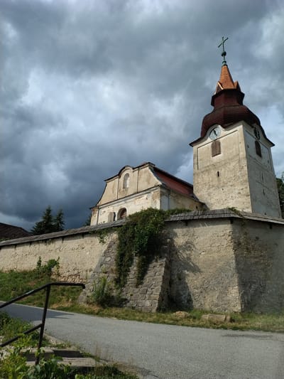 The image shows a stone church with a tower and a red-tiled roof, located in Hlávkov, Czechia. The church is surrounded by a stone wall, and there is vegetation growing on the wall. The sky is cloudy, with dark clouds in the background. A road runs in front of the church, and there is a metal railing on the left side of the road. The church tower has a clock on it. The overall scene appears to be one of an old, historic religious building in a rural setting.