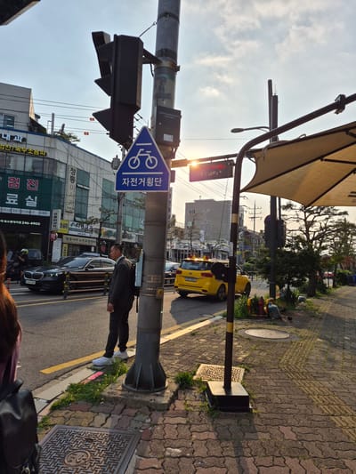 This image captures a street scene in Suwon-si, South Korea, with a focus on a traffic signal pole and its immediate surroundings. The sun is low in the sky, casting long shadows and a warm glow, suggesting it is either early morning or late afternoon.
Prominently featured is a traffic light pole with multiple signals, including what appears to be a bike lane signal with a blue triangular sign depicting a bicycle. Below this sign are Korean characters, "자전거횡단" which translates to "bicycle crossing". A man in a dark suit and white sneakers stands near the pole, looking to his left.
The street is paved with asphalt, marked with a yellow line. A black sedan is parked partially on the road, and a yellow taxi is visible further down the street. In the background, several buildings, characteristic of an urban environment, are present, with shops on the ground floors. Power lines are strung overhead.
The sidewalk is paved with interlocking brick pavers and features a textured surface for tactile guidance. A large, fabric sunshade extends over part of the sidewalk, supported by poles. There is also vegetation growing in patches along the sidewalk and around the base of the pole. A manhole cover is visible on the ground near the pole. The overall atmosphere is that of a typical urban street during a time of golden hour.