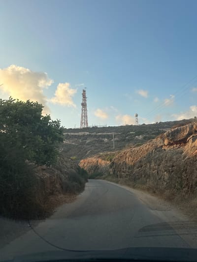 The image depicts a paved road winding through a rocky, arid landscape under a clear blue sky with scattered clouds. A large red and white communication tower stands prominently on a hill in the distance, accompanied by another smaller, domed structure on a separate tower further along the ridge. Electrical wires are visible strung between poles, spanning across the hillside. Lush green foliage lines the left side of the road, contrasting with the dry, rocky terrain on the right. The scene appears to be a remote, possibly mountainous area within the Mate Asher Regional Council, Israel, likely at dawn or dusk given the warm light on the clouds. There are no people visible in the image.