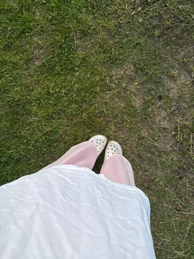 The image shows a close-up, overhead view of a person's lower body, from the waist down, as they stand on a grassy area. The person is wearing a white t-shirt or dress that drapes over their legs and a pair of white Crocs. The clothing is a pale pink color. The ground is covered with green grass and some patches of dry earth and fallen leaves. The setting appears to be outdoors, possibly in a park or yard, in the city of Ivatsevichy, Belarus.