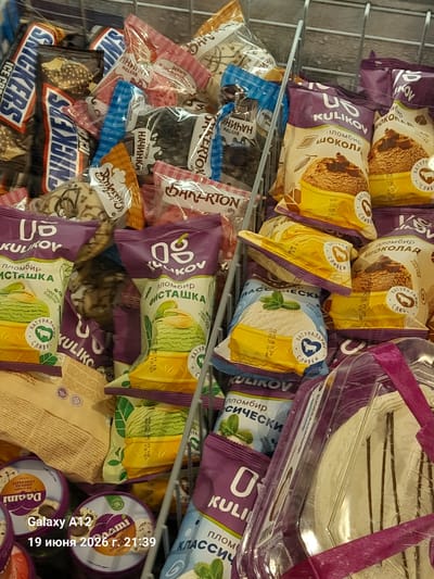 The image shows an assortment of individually wrapped frozen desserts, likely ice cream bars and cones, in a retail display. Many of the packages feature branding from "KULIKOV". The packaging is primarily in shades of purple, yellow, and light blue, with some red and white accents. Visible brands include "SNICKERS" on some blue and white wrappers. Some packages depict images of the dessert inside, such as chocolate ice cream, pistachio ice cream, and what appears to be a white ice cream with mint. The desserts are arranged in a metal bin or rack. In the lower left corner of the image, a smartphone overlay displays "Galaxy A12" and the date "19 июня 2026 г. 21:39" (June 19, 2026, 9:39 PM), suggesting the photo was taken with this device at this time. The environment appears to be a supermarket freezer aisle or similar retail setting. There are no people visible in the image. The overall scene is a close-up view of a frozen food display.