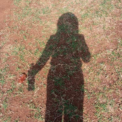A distinct, dark human-like shadow is cast centrally upon a dry, reddish-brown ground surface in Kisa Central ward, Kenya. The ground is sparsely covered with patches of green and desiccated grass, exhibiting some soil cracks. The shadow outlines a person facing away from the camera, with the left arm extending downward. Near the shadow's left hand, an irregular, brownish-red object, consistent with organic debris, lies on the soil. The right arm of the shadow is bent at the elbow, with the hand near the chest area. Direct sunlight strongly illuminates the scene, producing the sharp silhouette.