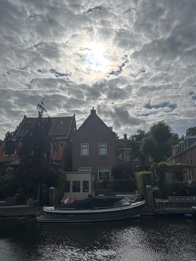 The image depicts a canal scene in Katwijk, Netherlands, under a partially cloudy sky where the sun is visible through breaks in the clouds. The foreground is dominated by dark, rippling water reflecting the sky and surrounding structures.

Along the canal bank, a small, dark-colored boat with a cover is moored. The boat has a registration number visible on its side: "85-38-YR". Adjacent to the boat is a wooden dock with metal ladders leading into the water. Further back, a building with a light-colored facade and windows is partially obscured by vegetation.

Behind the canal and dock, several houses are visible. One prominent house is made of brick with a pitched roof, featuring two windows on its upper floor and a chimney. Another house to its left has a red-tiled roof with solar panels installed. Dense green trees and lush foliage are interspersed between the houses, creating a suburban or semi-rural atmosphere. The overall impression is a tranquil waterside environment in a residential area of Katwijk.