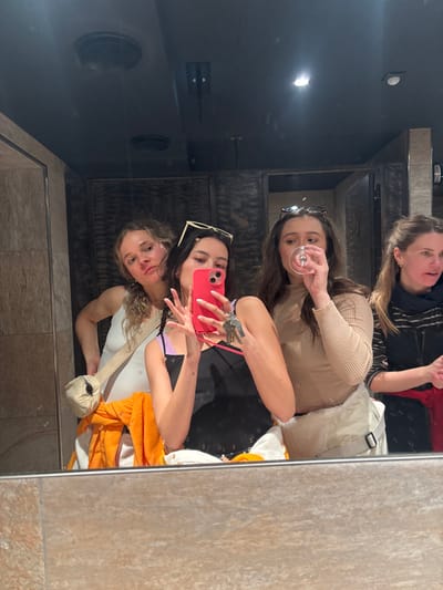 Four women pose for a mirror selfie in what appears to be a bathroom. The woman holding the phone wears sunglasses on her head and holds keys in her hand. Another woman holds a wine glass, and a third is partially visible in the top right. The reflection shows dark ceiling with a vent and a light fixture. The wall has tile, and a partially visible orange towel and bag appear in the reflection.