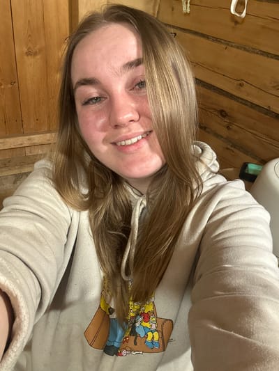 A young woman with blonde hair is smiling at the camera. She is wearing a light-colored hooded sweatshirt with a graphic of characters from The Simpsons on the front. The background appears to be wooden walls or paneling. There is no information in the image indicating the location is Bashur, Russia.
