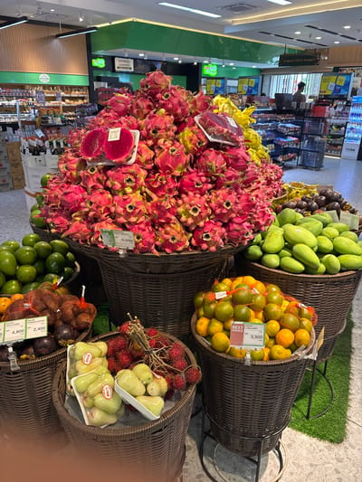 This indoor photograph captures a fruit display in a supermarket, likely located in Kerobokan Kelod, Indonesia, given the tags and currency on the products. Various wicker baskets overflow with fruits like dragon fruit, mangos, oranges, rambutan, and "jenggo" (local fresh fruit) alongside salak gula pasir and salak pondoh super. The dragon fruit basket is the most prominent and located centrally. Price tags are visible on many baskets. Aisles of supermarket products, a checkout counter, and other shoppers are in the background, providing context for the produce section within the store. The lighting is bright and general.