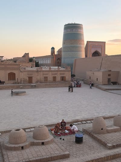 This image captures a scene within the Ichon-Qala, the walled inner city of Khiva, Uzbekistan, likely at dusk given the warm, soft light. In the foreground, two large, rounded clay ovens, possibly tandoors, sit on a brick base. To the right of these ovens, a group of people are gathered on a rug, some appearing to be eating or preparing food. A blue and white mosaic-patterned bin is also present.

The midground reveals a wide, paved courtyard. On the left side of the courtyard, a woman walks near a circular well structure. Further into the courtyard, a group of people are congregated around what appears to be a market stall selling various items. Several individuals are also seen walking or standing in various parts of the courtyard.

Dominating the background is the Kalta Minor Minaret, a distinctive, unfinished turquoise-tiled minaret known for its cylindrical shape and intricate geometric patterns. Adjacent to it is the Madrasah of Mohammed Rahim Khan, characterized by its imposing walls and decorative facade. Other traditional brick buildings with arched windows and doorways are visible, contributing to the historic architectural landscape of Khiva. The sky above is a pale gradient of blue and soft orange, indicating either sunrise or sunset.