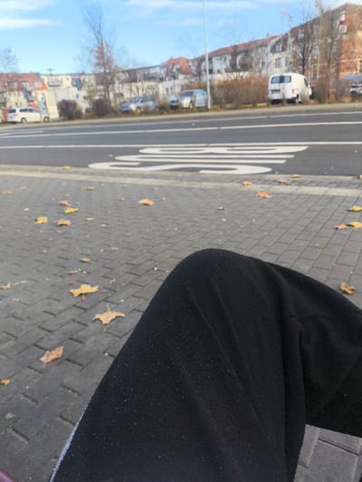 This image, taken in Freital, Germany, presents an outdoor scene from a low, first-person perspective. In the immediate foreground, the knees and thighs of an individual, clothed in black fabric trousers with visible lint, are partially visible in the lower right section. The subject's legs rest above a light gray paved sidewalk composed of rectangular interlocking stones. Scattered yellow-brown deciduous leaves are dispersed across the sidewalk surface.

Immediately beyond the sidewalk, a dark asphalt road runs horizontally across the frame. White road markings are distinctly visible, including a prominent number "30" indicating a speed limit, accompanied by parallel dashed lines. On the far side of the road, a light pole stands vertically. Behind the roadway, a strip of vegetation consisting of bare deciduous trees and dormant shrubs separates the street from a parking area.

Several vehicles are parked along this strip, including a white panel van, a white SUV, and at least two silver-colored passenger cars. In the background, multiple multi-story residential buildings with red-tiled roofs are discernible. The sky above is clear and light blue with minimal cloud presence. The scene is illuminated by daylight, suggesting autumn or early winter due to the bare trees and fallen leaves. No other individuals are present or engaged in discernible actions within the visible frame.