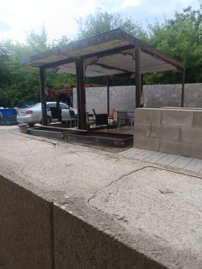 The image shows an outdoor scene in Milovice, Czechia. In the foreground, a rough, textured concrete wall runs diagonally across the frame, with a sharp edge in the center of the image. Behind the wall, a dilapidated open-air structure with a rusted metal frame and torn roofing material stands. Underneath this structure, a silver sedan with its doors open is parked. To the right of the structure, a wall made of concrete blocks is visible, and further in the background, trees and a cloudy sky can be seen. The overall impression is one of disrepair and abandonment.