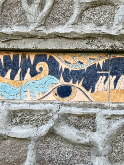 A close-up, eye-level, outdoor, medium shot shows a section of a textured stone wall with a decorative tile inlay. The wall is constructed of irregular, rough-hewn grey stones of various sizes, interspersed with cement mortar. The mortar is cracked and appears weathered. The tile inlay is a horizontal band, approximately 20cm high, featuring a stylized, abstract design with a large, prominent eye in the center. The eye has a dark blue pupil and is surrounded by pale yellow and blue wavy lines, possibly representing water or a dynamic element. Above the eye, the design continues with jagged yellow shapes and a dark blue background with fine speckles, reminiscent of teeth or a monstrous mouth. To the left of the eye, there are white, hand-like shapes emerging from the blue and yellow elements. To the right, there are more white, stylized shapes. The tile inlay appears to be part of the facade of a building located in Sinemorets, Bulgaria.