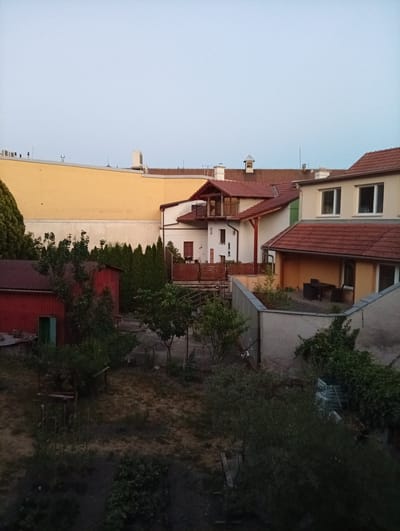 This is a medium-angle, eye-level, exterior, full, daylight shot of a residential backyard in Tišnov, Czechia. The scene depicts a backyard area with several houses and sheds, surrounded by trees and vegetation. In the foreground, a red shed with a green door is visible. Behind it, a fence separates the yard from other properties. Several trees, including evergreens and deciduous trees, are scattered throughout the yard. In the background, the scene shows the rear of houses with red tiled roofs. The sky is a pale blue, suggesting early morning or late evening. There are no people or animals visible in the image.