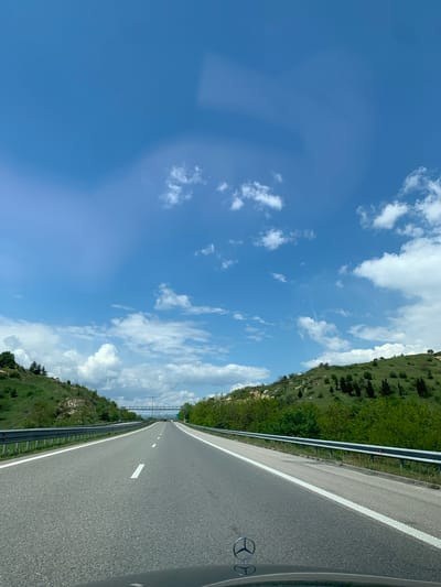 The image captures a road trip perspective, looking forward from inside a vehicle on a bright, clear day. The original photo appears to be rotated 90 degrees counter-clockwise, showing the horizon vertically.
When viewed correctly, the scene features a gray asphalt road stretching into the distance on the right side of the frame, marked with a solid white line along its shoulder and dashed white lines for lanes further ahead. A silver guardrail parallels the road on the right. To the left, a verdant hillside covered in green grass and scattered trees rises. The upper portion of the image is dominated by a bright blue sky filled with numerous fluffy white clouds.
The activity depicted is traveling or driving on a highway or country road through a scenic, hilly, or mountainous region. The weather is sunny and clear, suggesting it's likely midday or early afternoon due to the strong natural light. A notable detail is the partial view of a Mercedes-Benz logo (the three-pointed star within a circle) visible in the bottom right corner, indicating the make of the vehicle from which the photo was taken. The location context provided is Marikostinovo, Bulgaria, which aligns with the rural highway and green landscape seen. There is no other legible text visible in the image.