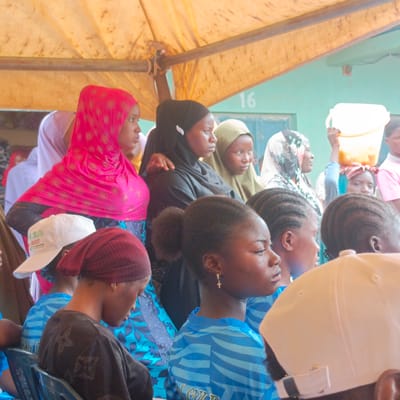A group of people, mostly women and girls, are gathered outdoors under a yellow canopy in Gudi, Nigeria. The location appears to be near a building, indicated by the number "16" on its exterior. Many of the women are wearing headscarves, including pink, black, green, and white patterned scarves. Some women are in blue striped shirts. A girl in the center of the image has braided hair and is wearing a cross earring. Another person in the foreground wears a white baseball cap. A woman stands with a white bucket on her head in the background.