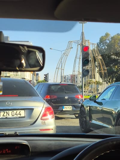 The image displays a stationary traffic scene observed from inside a vehicle through its windshield in Gonyeli, Northern Cyprus / Cyprus, under clear daylight conditions.

In the immediate foreground, the top of a vehicle dashboard is visible, showing a digital display indicating "210 km" and "27°C." A portion of a dark steering wheel rim is present on the lower right, and the rearview mirror is positioned at the top center, reflecting an indistinct interior.

Ahead, three vehicles are stopped in traffic. Directly in front, a silver-grey Mercedes-Benz sedan (license plate TZN 044) occupies the left lane. A small sticker with partially legible text is affixed to its rear bumper. Behind the Mercedes, in the same lane, a dark grey Volkswagen Golf hatchback (license plate PV 713) is stopped, with its rear brake lights illuminated. To the right of the Volkswagen, in an adjacent lane, the front left quarter, wheel, and side mirror of a dark green Porsche Taycan electric sedan are visible.

A prominent traffic light pole stands centrally, featuring a vertically oriented signal head with the top red light illuminated. This pole is encased within a large, ornate, gold-colored metal decorative framework that arches outwards. In the background, several multi-story buildings line the street, and green foliage is visible on the right. The sky is clear blue.