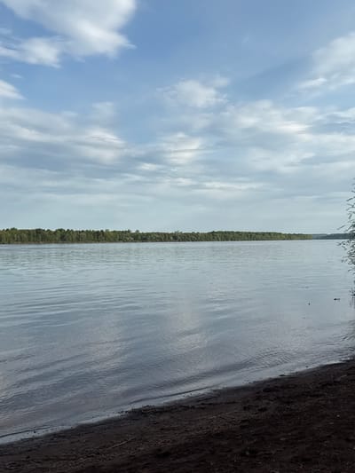 The image depicts a body of water, likely a river or lake, in what appears to be a natural setting. The left side of the image shows a blue sky with scattered white clouds. A narrow strip of trees lines the center of the image, separating the sky from the water. The water surface reflects the sky, creating a mirrored effect. The right side of the image shows the edge of the water meeting the shoreline, which consists of dark soil or mud. The photograph is taken from an angle that is rotated clockwise. The presence of trees and the body of water suggest a location outside of a densely populated urban area, possibly the Doksha area of Russia. No people or actions are visible in the image.