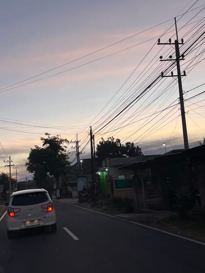 This media file captures a street scene at dusk in Purworejo, Indonesia. The image is oriented vertically, with the sky occupying the left portion and the road and buildings on the right. A white car with its taillights illuminated is driving away from the viewer on the right side of the road. The car's license plate is visible and reads "W 1850 YP". Overhead, a dense network of power lines crisscrosses the sky, supported by utility poles. The sky itself displays a gradient of colors, transitioning from a soft purple and pink near the horizon to a pale yellow and gray higher up, indicating the twilight hour.
On the right side of the road, buildings and trees are visible, partially obscured by the dim light. Some of the buildings appear to be residences or small shops, with some lights on inside. A motorcycle is parked in front of one of the buildings. The overall atmosphere is calm and quiet, typical of the early evening in a residential or semi-urban area. The presence of the power lines and the car suggests a functional, inhabited location. The weather appears to be clear, allowing for the visibility of the sunset colors.