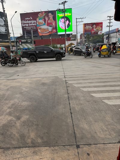 This image shows a street scene in Dagupan City, Philippines, with multiple vehicles and billboards. In the foreground, a pedestrian crossing is visible on a concrete road. A black pickup truck is stopped at the crossing. Several motorcycles, some with passengers, are on the road. Yellow tricycles, a common mode of transportation in the Philippines, are also present. In the background, large billboards advertise Jollibee, Chowking, and another unspecified product. The sky is overcast, suggesting it might be daytime. The overall impression is a busy urban environment with diverse modes of transportation and prominent advertising.