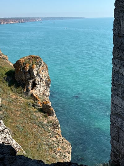 This is an outdoor shot of a rocky cliffside overlooking a body of turquoise water, likely the Black Sea. The vantage point is from within a stone structure, possibly a fortress or castle ruins, framing the scene.
The cliff face is covered with vegetation, including grass, scrub, and wildflowers. A distinct rock formation protrudes from the cliff, displaying a mix of grey and orange tones.
In the background, across the water, a distant coastline is visible. Two birds are seen flying over the water, adding a sense of scale and movement. The water itself has subtle wave patterns and variations in color.
Given the city of Balgarevo, Bulgaria as the location, this scene likely depicts the cliffs along the Black Sea coast near the Kaliakra Fortress.