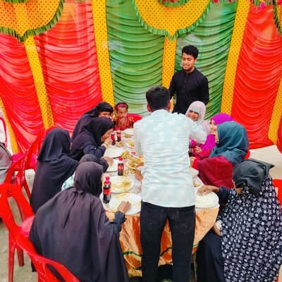 The scene in Lalmohan, Bangladesh, depicts approximately ten individuals gathered around a rectangular table under a temporary canopy. The canopy's interior is decorated with vertical drapes of red, green, and yellow fabric. A man in a light blue patterned shirt stands with his back to the camera, positioned behind a gold-colored tablecloth-covered table, seemingly serving or attending to the seated guests. Another man in a black long-sleeved shirt stands behind the table, smiling. Eight individuals, predominantly women, are seated on red plastic chairs around the table. Several women are dressed in black hijabs or niqabs, while others wear colorful head coverings and clothing. A young girl with a red headband is also seated. White plates containing rice and various curry-like dishes, along with multiple Coca-Cola bottles and smaller red bottles, are spread across the table. Seated individuals are actively eating or reaching for food. The ground surface is light-colored.