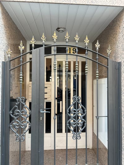 This is a medium shot, eye-level, outdoor photograph of a gated entrance to a building. The gate is made of dark gray metal bars, with decorative elements on top and in the center of each panel. The gate is open, revealing a white door with glass panels behind it. To the left of the door are a set of mailboxes. Above the door, a house number "19" is visible in black numerals on a gold background. The building's exterior walls are covered in a speckled, brown and pink granite-like material. The ceiling above the entrance is made of white, horizontal planks. The scene appears to be in the city of Castelló De La Plana, Spain, based on visual cues of architectural style and materials common in the region. No people are visible in the image.