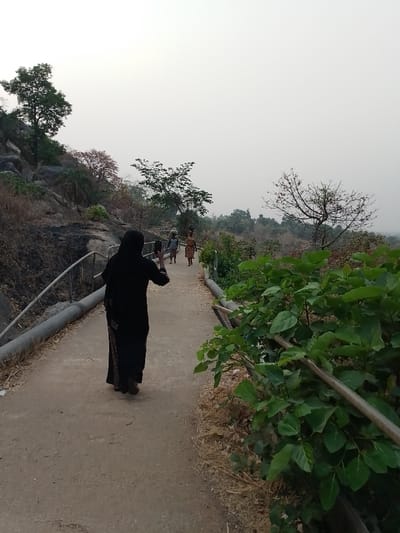 A woman in a black robe stands on a concrete path, holding a phone as if taking a photo. She faces away from the viewer, looking towards two women carrying items on their heads in the distance. The path is bordered by railings and greenery on the right, and a rocky slope with trees on the left. The sky is overcast, suggesting either early morning or late evening.