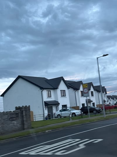 The image depicts a street scene in Rathvilly, Ireland, on a cloudy day. In the foreground, a grey asphalt road is visible with white markings. Beyond the road, a row of modern white houses with dark tiled roofs lines the street. Several cars are parked in front of the houses, including a white sedan, a black SUV, and a red car. A black metal fence separates the properties from the road. To the left, a stone wall with a castle-like crenellated top is situated in front of one of the houses. A street light stands tall on the right side of the image. The sky is overcast with heavy grey clouds, indicating a possibly inclement weather. No people are visible in the scene.