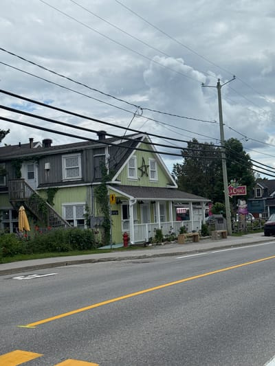 The image depicts a street view in Chelsea, Canada, under a cloudy sky. The foreground shows a road with a yellow dividing line and yellow pedestrian crossing markings. To the right of the road, a sidewalk runs along the front of a building complex. The building on the left is painted light green and has an exterior wooden staircase leading to the upper level. A yellow umbrella is visible in front of the building, accompanied by greenery and red flowers.

A prominent feature is a sign pointing to "La Cigale," which appears to be an ice cream shop, indicated by a large, colorful ice cream cone statue next to the sign. The building associated with "La Cigale" has a white porch with railings and wooden benches placed outside. Further to the right, another building, dark blue in color, is partially visible. A red fire hydrant stands near the corner of the green building. Overhead, a dense network of power lines crisscrosses the sky. The overall scene suggests a commercial area with residential elements in a town setting.