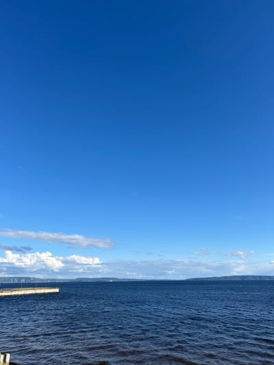 The image depicts a wide, dark blue river under a bright, clear sky with scattered white clouds. On the left side of the frame, a long, light-colored pier extends into the water. The pier has railings and appears to be made of concrete or a similar material. In the distance, across the river, a faint, low-lying shoreline with a slightly hilly landscape is visible. The water is somewhat choppy, with visible ripples and waves. The lower right corner of the image shows a small portion of the riverbank with some dry vegetation. The overall impression is a serene natural landscape with a man-made structure. The location is identified as Podstepki, Russia. There are no people or animals visible in the image.