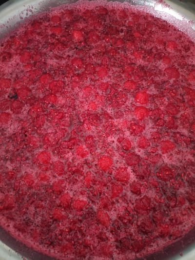 This is a close-up, overhead shot of a large quantity of raspberries being cooked in a metal pot. The raspberries are a deep red color and are surrounded by a pinkish froth, indicating they are actively boiling. Some of the raspberries appear whole, while others have broken down, releasing their juices. The texture is a mix of smooth liquid and chunky fruit pieces. The scene likely takes place in a kitchen or food preparation area in the city of Kyzyl Suu, Kyrgyzstan, where jam or preserves are being made. There are no people or specific environmental details visible beyond the pot and its contents.