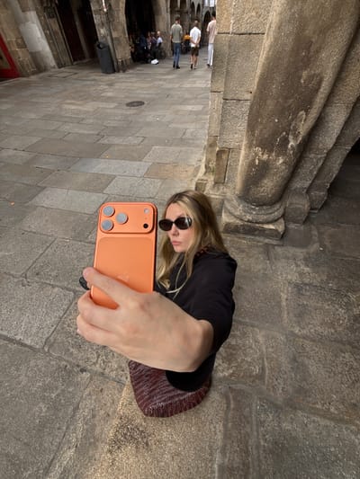 A woman is taking a selfie with an orange smartphone in an outdoor courtyard in Santiago de Compostela, Spain. The courtyard is paved with large, grey stone slabs, and there are stone arches and columns in the background. Several people are walking in the distance, and some are seated at tables in an outdoor cafe. The woman is wearing sunglasses and a black top. The smartphone has a triple-lens camera system.