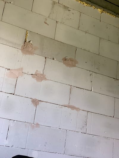 The image shows a section of a wall constructed from white, rectangular cinder blocks. The blocks are laid in a staggered pattern, with visible mortar lines between them. Several of the blocks have small, irregular patches of a light brown, powdery substance applied to their surfaces. In the upper portion of the image, yellow foam insulation is visible along the top edge of the wall. The overall scene appears to be an unfinished construction or renovation site. The image is a close-up of the wall and does not provide context for its location within Petrovskiy, Russia, or any specific actions or interactions occurring.
