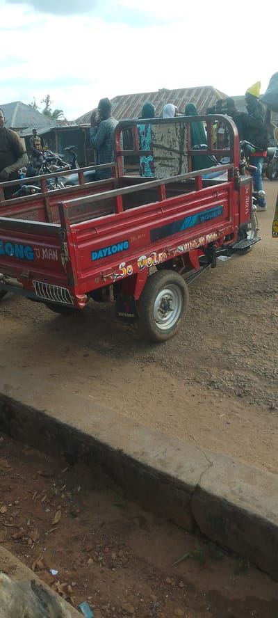 The image shows a red three-wheeled utility vehicle, commonly known as an "okada" or "keke," parked on a dirt road. The vehicle has "DAYLONG" branding visible on its side, along with other text that is partially obscured or difficult to read, such as "J'MAN" and "So DoIN." A mudguard on the rear wheel also displays the "DAYLONG" logo.
Several people are visible in the background. Some appear to be standing or sitting around the vehicle, while others are farther back near buildings with corrugated metal roofs, suggesting a rural or semi-urban setting. The sky is overcast, indicating a cloudy day. The overall scene depicts a typical street or roadside area in what appears to be a populated area in Doka-Tofa, Nigeria. The presence of multiple people and the utility vehicle suggests it might be a marketplace, a transportation hub, or a gathering point. The ground is uneven and covered with dirt and gravel. Some of the people are dressed in casual clothing, and one individual in the background is wearing a bright yellow cap. The image is taken from a slightly elevated perspective, looking down at the utility vehicle.