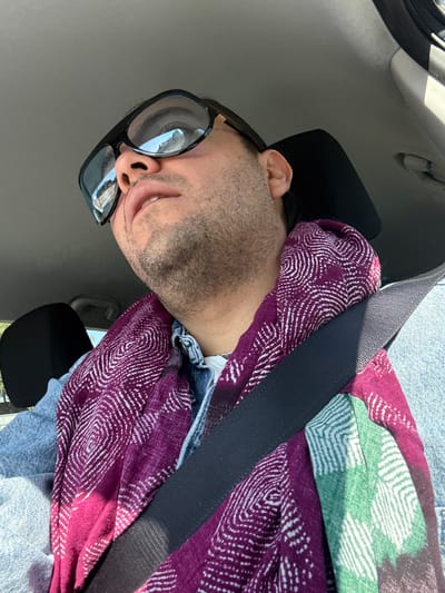This image, captured in the city of San Juan Cuautlancingo, Mexico, depicts an adult male seated within the passenger compartment of a vehicle. The individual is positioned from a low-angle perspective, facing slightly upward and to the right. The subject has light-to-medium skin tone with visible stubble on the jawline and chin, and dark hair on the visible side of the head. Black-framed sunglasses are worn, reflecting the vehicle's interior and external sky in their lenses. The person is dressed in a light blue denim shirt or jacket over a lighter garment visible at the neckline. A multi-patterned scarf, predominantly dark red/magenta with white concentric designs and a visible segment of green with similar white patterns, is wrapped around the neck and upper chest. A black vehicle seatbelt is securely fastened across the individual's torso, extending from the upper left to the lower right of the frame. The vehicle's headliner is light grey, and a dark-colored headrest is partially visible behind the subject's head. Natural light illuminates the scene, casting shadows on the scarf and seatbelt. The individual's mouth is slightly ajar, and the gaze is directed upward.