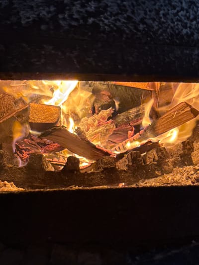 The image shows a close-up view of a fire burning in a fireplace in Kandry, Russia. The fireplace appears to be made of dark, textured material, possibly brick or stone. Inside, several pieces of wood are stacked and engulfed in flames. The fire is bright orange and yellow, with visible smoke and heat distortion. The wood pieces vary in size and shape, with some appearing rough and unfinished. The overall scene depicts a warm and active fire.