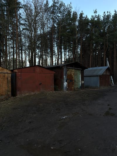 The image depicts a row of derelict garages situated in Lavriki, Russia. The garages are constructed from corrugated metal and wood, exhibiting signs of rust, decay, and weathering. A dense forest of tall, bare trees forms the backdrop. The ground in the foreground appears to be a dirt path or roadway, muddy and wet. The sky is overcast, suggesting a grey and damp day. There are no people visible in the scene.