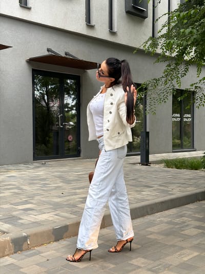 A person stands on a paved walkway in front of a modern building. The person is wearing a white denim jacket over a white top and light-colored jeans. They have on strappy high-heeled sandals with a leopard print pattern. Their long, dark hair is pulled back into a ponytail. They are looking down and to the side, and appear to be adjusting their sunglasses.
The building has a grey textured facade with dark-framed windows and a dark awning over a doorway. Green foliage is visible in the upper left corner, suggesting it's an outdoor setting. The walkway is made of grey rectangular pavers.
Reflections in the window show trees and an outdoor scene. Text is visible on the windows of the building, reading "JOIN& ENJOY" repeated. The lighting suggests it is daytime, possibly late afternoon due to the angle of the shadows and the warm light. The overall impression is casual and stylish.