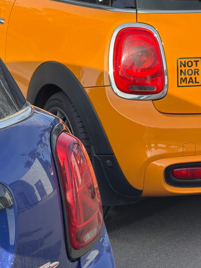 The image displays a close-up, oblique view of the rear sections of two cars parked bumper-to-bumper, likely on a street or in a parking area in Luga, Russia, during daytime. The scene is brightly lit by the sun, causing reflections on the vehicles' glossy surfaces.
The car on the left, partially in the background, is a vibrant yellow. Its distinctive red rectangular taillight, framed by a chrome trim, is prominent. Below the taillight, a black plastic rear bumper or fender flare and a portion of a tire are visible. On the upper rear panel of the yellow car, there is a black rectangular sticker with white text arranged vertically, which clearly reads "NOT NORMAL".
In the immediate foreground, partially overlapping the yellow car, is a blue car. Its metallic blue paint reflects light and shows distorted reflections of what appear to be trees or buildings. This car also features a red taillight with a black trim. A small part of a car badge on its trunk is visible, with the word "COOPER" partially legible, suggesting it might be a Mini Cooper. No people are visible, and the cars are stationary.