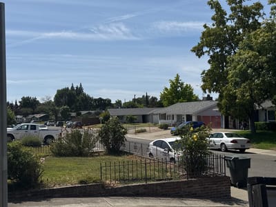 This daytime image depicts a tranquil suburban residential street in North Highlands, United States, under a clear, bright sky. The scene is dominated by a row of single-story houses on both sides of the paved street, set back from well-maintained front yards.
In the foreground, a low brick wall topped with a black wrought-iron fence separates a grassy lawn with several bushes from the sidewalk and street. On the left, a white pickup truck is parked, partially visible behind the fence. To the right, a white sedan, possibly an Acura, is parked on the street, its license plate unreadable. A large dark green garbage bin stands on the sidewalk next to the white sedan. Further down the street, a blue car is parked in front of a house with an open garage, revealing stacked items inside. Other vehicles are visible in the distance.
The landscape is rich with vegetation, including several large, leafy green trees that provide ample shade, particularly on the right side of the frame and in the background. Bushes, some with yellow flowers, are scattered throughout the yards. Utility poles and power lines are visible above the rooftops in the mid-ground.
The sky is a pale blue with scattered, wispy white clouds and faint contrails, suggesting fair weather. The bright sunlight casts distinct shadows, indicating a clear, sunny day. No people are visible, and no specific activities are taking place, portraying a quiet, everyday street scene.