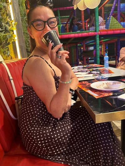 The image shows a woman with dark hair and glasses drinking from a black cup. She is wearing a dark polka-dot dress and is sitting in a red booth. In the background, a colorful indoor playground is visible. The scene appears to be in a restaurant or entertainment venue, possibly a family-friendly establishment. The woman is the primary subject, with the playground serving as a secondary element of the environment. There are no obvious indicators that place this scene specifically in Carretera De San Martín, Spain.