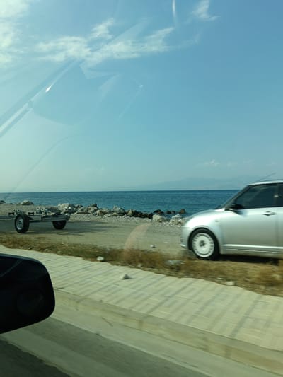 The image captures a scene from within a vehicle traveling along a coastal road. The foreground shows a portion of the vehicle's interior, including a side mirror and a reflection of the sky and clouds on the windshield.
The exterior scene reveals a clear blue sky with scattered clouds. The sea stretches out to the horizon, meeting a distant mountain range. The coastline is rocky, with a mix of sand and pebbles. In the mid-ground, a silver car is driving on the road. To the left of the car, a boat trailer is visible on the side of the road. The road itself is paved with interlocking bricks, and there is a strip of dry grass and vegetation bordering it. The overall impression is a scenic drive along the coast of the Municipal Unit of Sikyona, Greece.