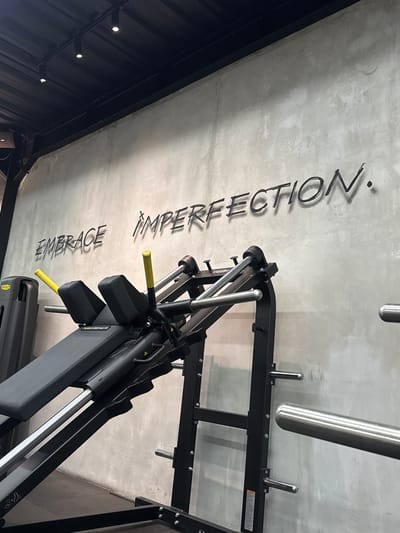 The image shows the interior of a fitness facility in Pererenan, Indonesia. A black squat rack machine is in the foreground. A concrete ceiling is visible with the words "EMBRACE IMPERFECTION." written on it in black. The lighting in the facility is dim.