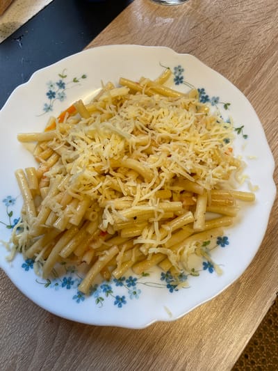 A white ceramic plate with a blue floral pattern is filled with cooked pasta, topped with shredded cheese. The plate sits on a light brown wooden table. The scene is indoors.