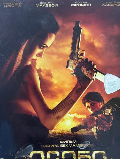 This image is a movie poster, featuring Angelina Jolie prominently. She is lying on her side, holding a silver-colored handgun above her head. Her arm has various tattoos, including "TRARS," a numerical sequence of "0101," and the Roman numeral "XIII." Another figure, a man, is partially visible in the upper right corner, also holding a gun. A "DVD Video" sticker is affixed to the poster. The background appears to be fiery and smoky. The poster also includes names of actors and the director in a Cyrillic alphabet. The words “АНДЖЕЛИНА Джоли”, “ДЖЕЙМС МАКЭВОЙ”, “МОРГАН ФРИМЭН”, and “КОНСТАНТ ХАБЕНС” are displayed. There are no obvious cues that the image was captured in Chuprino, Russia.