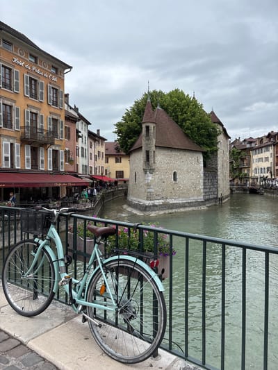 The image shows a light blue bicycle parked on a pedestrian walkway next to a railing. The walkway overlooks a canal with murky green water. On the opposite side of the canal are buildings with colorful facades and balconies. A prominent stone building with a red-tiled roof and turrets sits on a small island in the canal. The sky is overcast. The area appears to be a historic district in Annecy, France, likely the Palais de l'Isle area, a famous medieval structure located in the center of the Thiou canal. The bicycle is a modern hybrid model with a basket and a saddlebag. There are potted flowers on the railing. In the background, people can be seen walking along the canal and sitting at outdoor cafes. The architecture suggests a European city with a rich history.