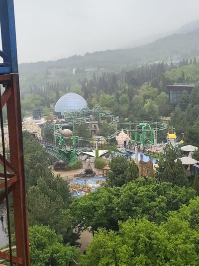 The image depicts an overview of a portion of a theme park located in Симеизский поселковый совет, Ukraine. A prominent feature is a geodesic dome, possibly a planetarium or a themed attraction, situated near a green-painted roller coaster. The coaster snakes through the park's landscape, which is heavily vegetated with mature trees. Several themed structures and attractions are visible, including what appears to be a water ride with boats. An observation tower provides an elevated vantage point. In the lower left corner, a person can be seen walking along a paved path. The weather appears overcast, with a hazy or foggy background.