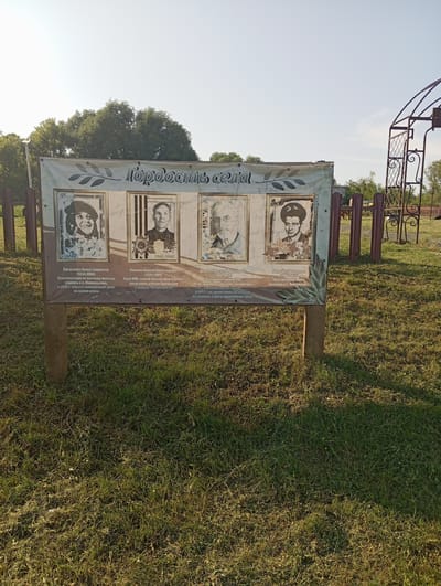 This is a full-frame, eye-level, outdoor shot of a memorial display board in the village of NovoKopylövo, Russia. The board, mounted on two wooden posts, features the title "Гордость села" (Pride of the Village) and displays four portraits with accompanying text. The portraits are of individuals, each in a framed section. The environment is a grassy area with trees and a metal archway visible in the background. The lighting suggests it is daytime, possibly morning or late afternoon due to the long shadows.